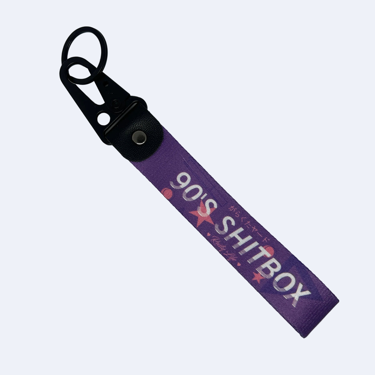 A purple fabric keychain strap with a black metal clasp, featuring the text '90'S SH*TBOX' in white and pink star graphics, displayed on a light blue background.