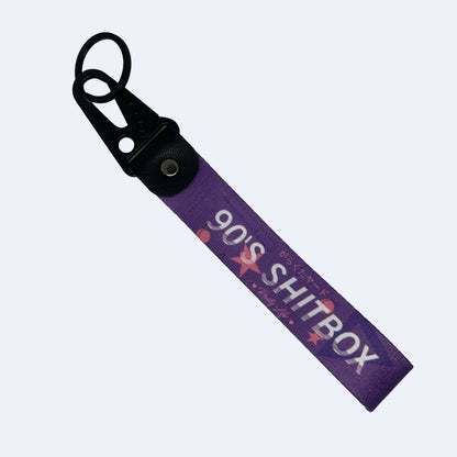 A purple fabric keychain strap with a black metal clasp, featuring the text '90'S SH*TBOX' in white and pink star graphics, displayed on a light blue background.