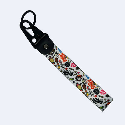 A fabric wristlet strap keychain featuring a white background with a colorful cartoon doodle pattern and the word 'BANG' repeated, attached to a black plastic carabiner clip with a silver metal snap button.