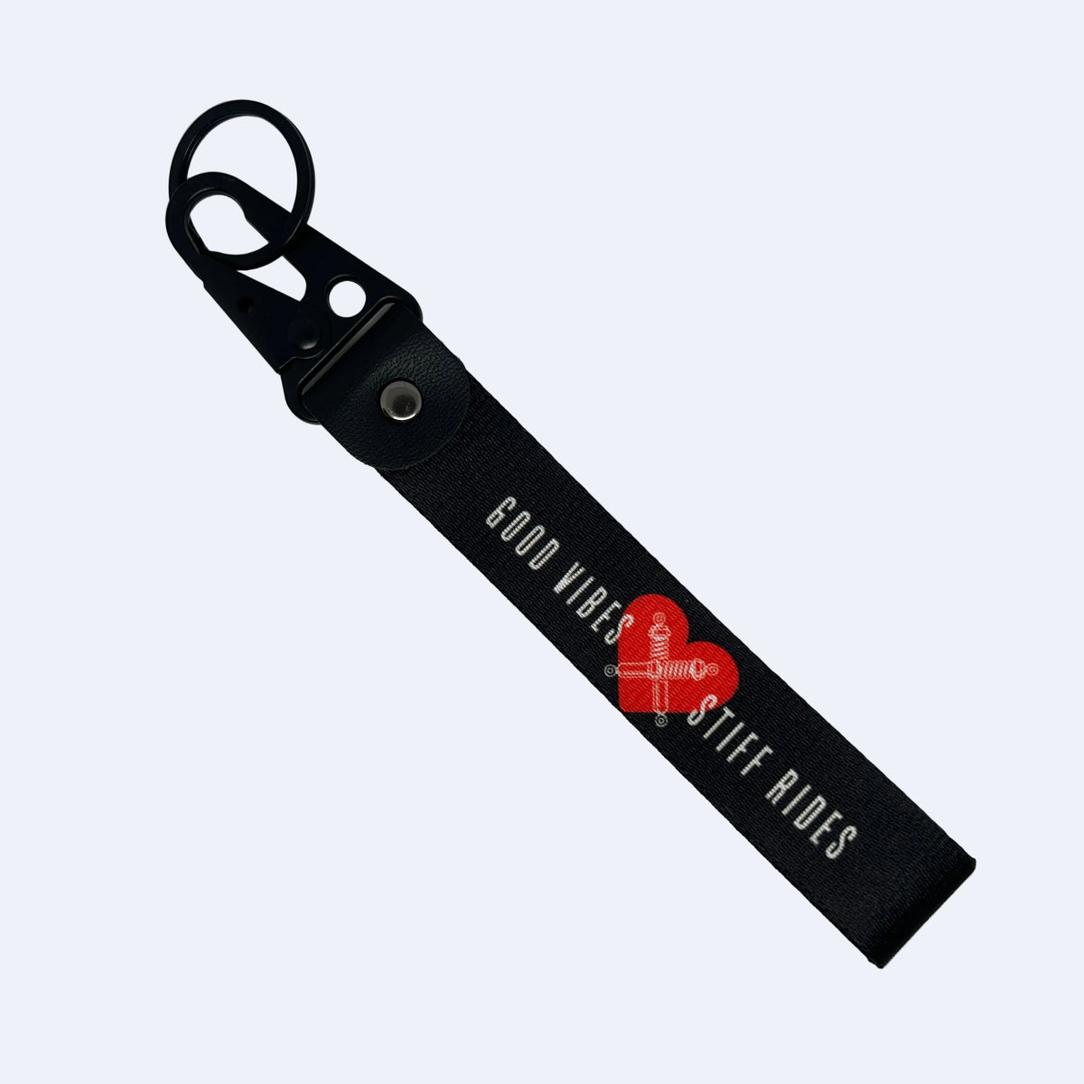A black leather keychain strap with a metal key ring and snap hook, featuring the text 'GOOD VIBES' and 'STIFF RIDES' in white, separated by a red heart graphic with a cross in the center. The keychain is photographed diagonally on a plain white background.