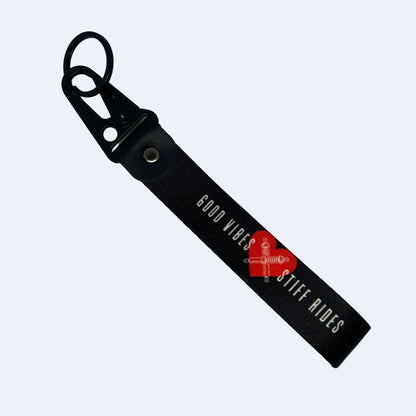 A black leather keychain strap with a black metal carabiner clip, featuring the text 'GOOD VIBES STIFF RIDES' in white. A red heart-shaped watermark is overlaid on the center of the strap.