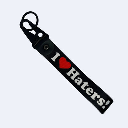 A black keychain strap featuring the text 'I' followed by a red heart and 'Haters!' in white, attached to a black metal key ring on a plain light gray background.