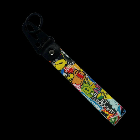 A fabric wristlet strap with a black faux-leather carabiner clip, featuring a colorful graffiti-style collage print with cartoon characters and text, shown on a light gray background.