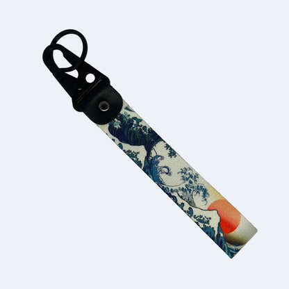 A canvas keychain strap featuring a print of Hokusai's 'The Great Wave off Kanagawa' in blue, white, and red, attached to a black plastic carabiner and black leather loop on a light gray background.