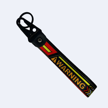 A black rubber strap keychain with a metal key ring and snap button, featuring a yellow and red warning graphic with the word 'WARNING' in yellow on a red background.